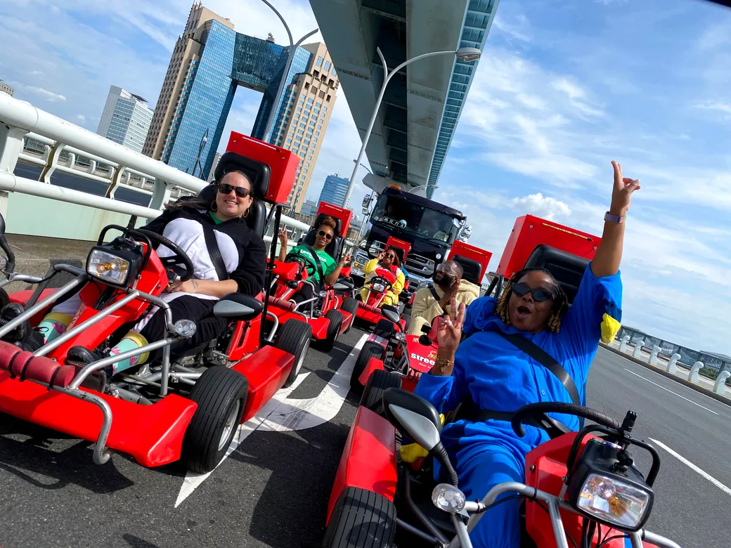 Group of people riding red go-karts on a bridge, smiling and posing, under a clear blue sky with city buildings in the background.