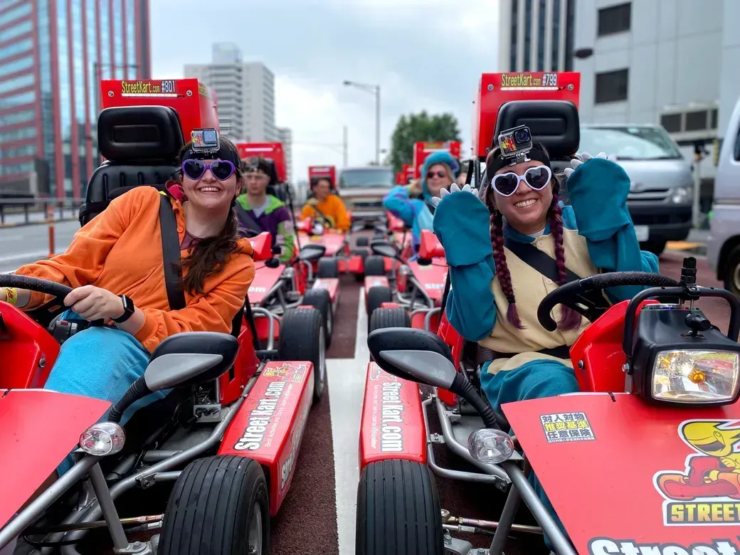 Two women in colorful outfits on red go-karts in a street parade, wearing playful sunglasses and smiling at the camera.