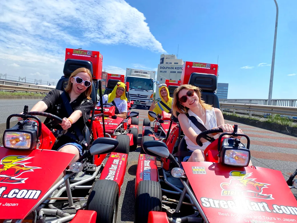 Group of people riding bright red go-karts on a sunny highway, smiles and blue sky above.
